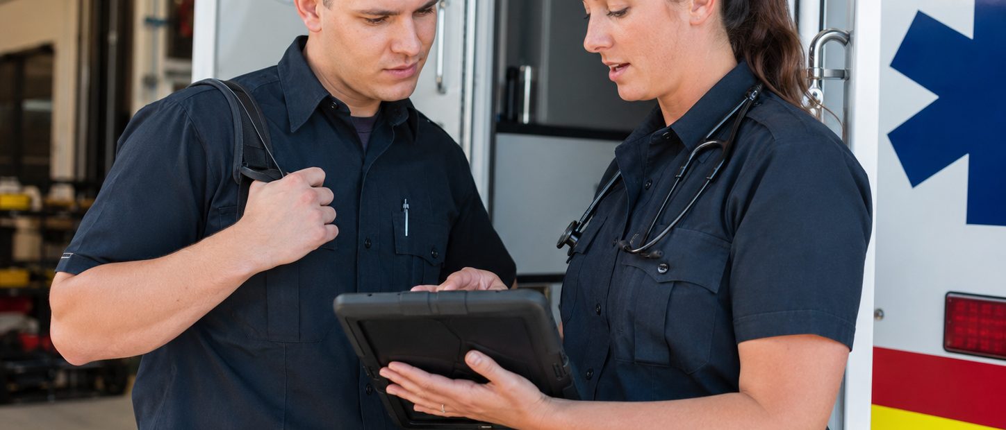 An EMS leader and field clinician review a tablet beside an ambulance during a training or evaluation discussion.